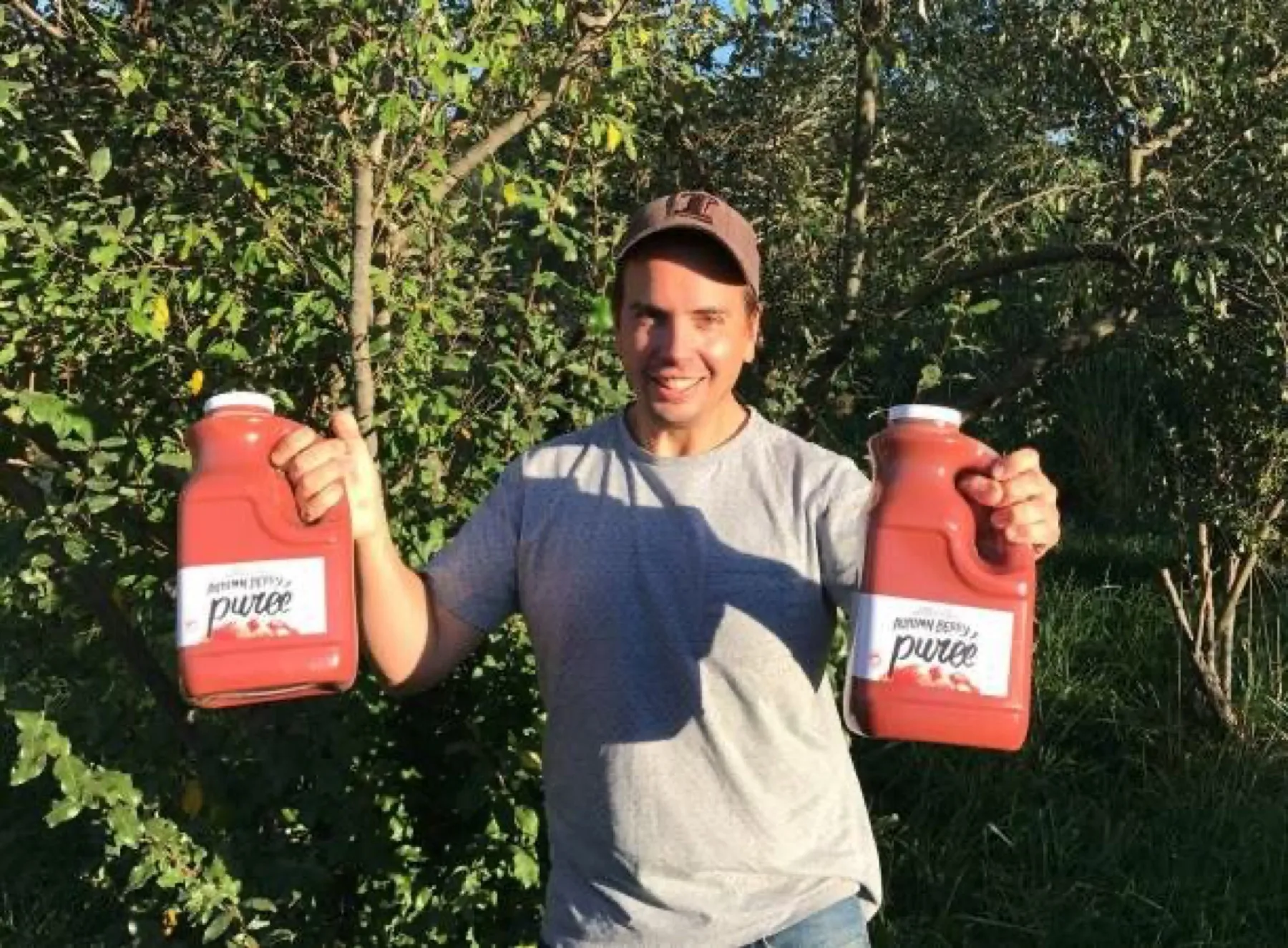 Dustin Kelly standing among autumn olive shrubs, holding up a jug of autumn berry puree in each hand.