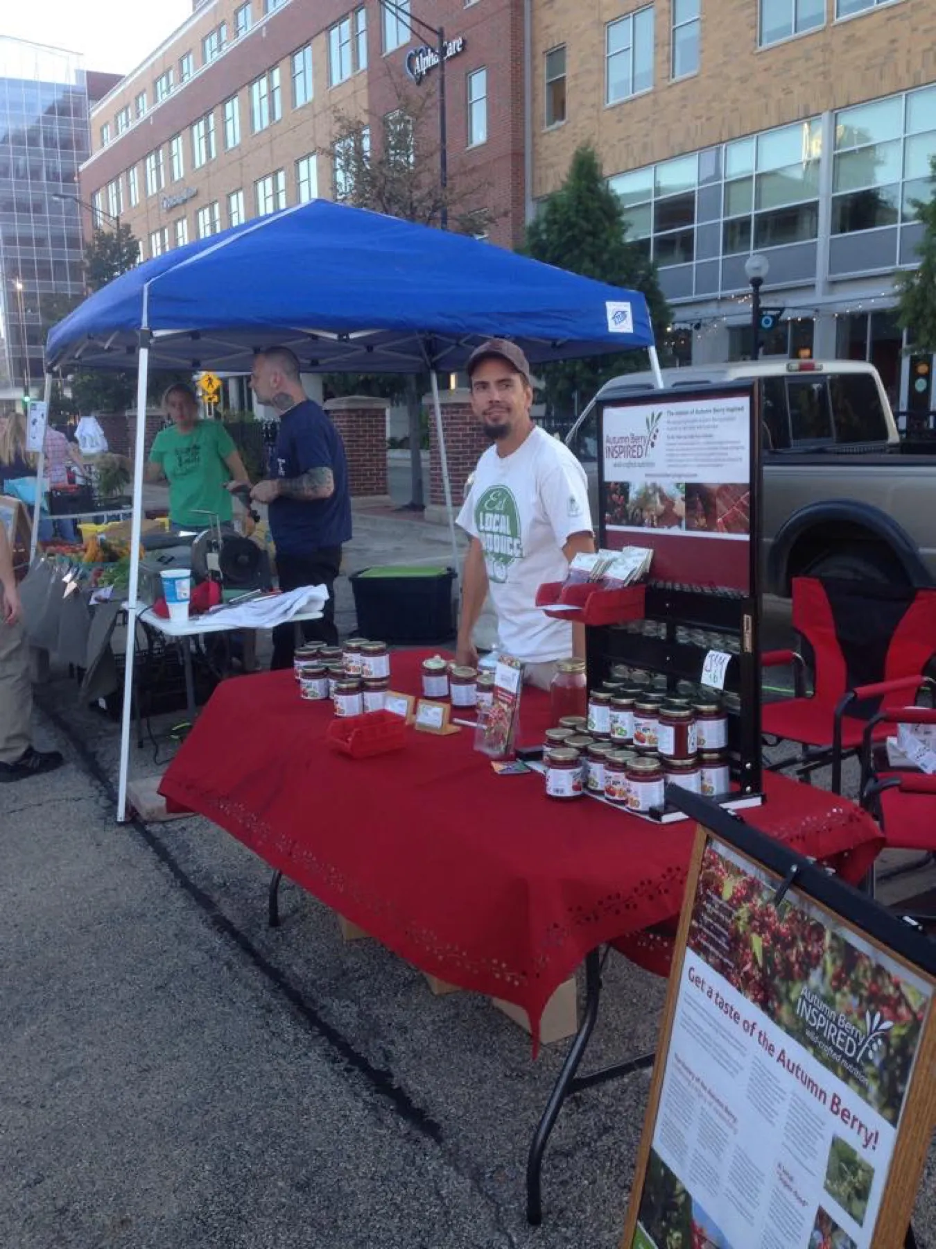 A farmers market stall under a blue canopy, with jars of jam on a red tablecloth.